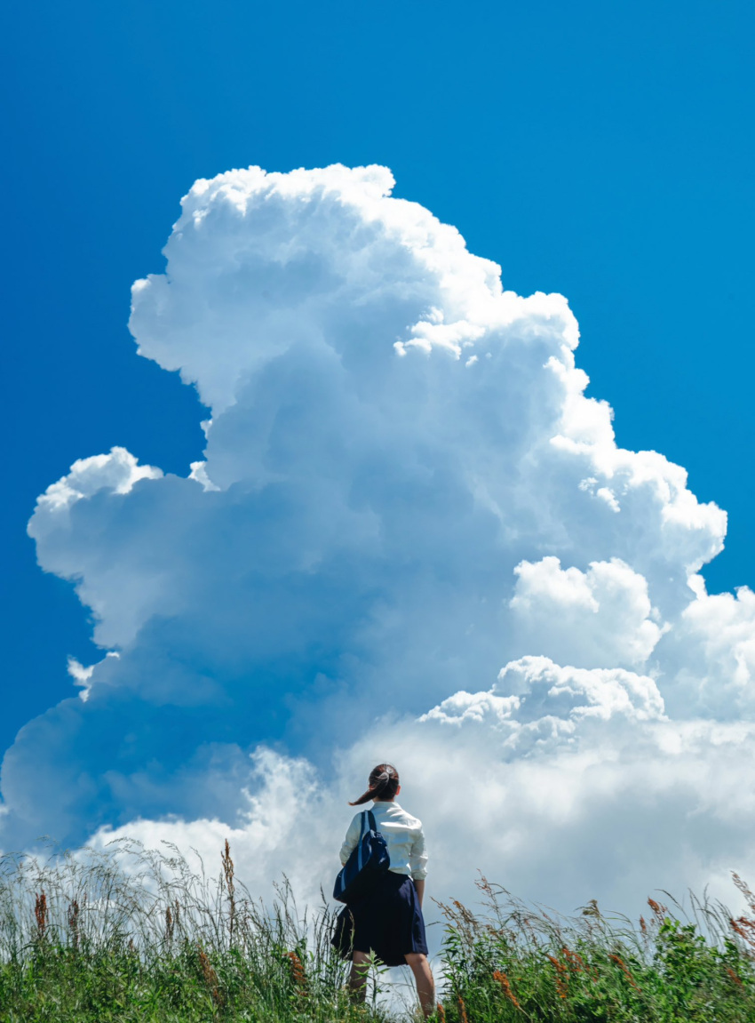 1girl bag black_hair blue_bag blue_skirt cloud cloudy_sky commentary_request cosplay_photo cumulonimbus_cloud day floating_hair from_behind grass highres iwato_suzume long_hair long_sleeves outdoors photo_(medium) pleated_skirt ponytail scenery school_bag school_uniform shirt shoulder_bag skirt sky solo standing suzume_no_tojimari tomosaki white_shirt