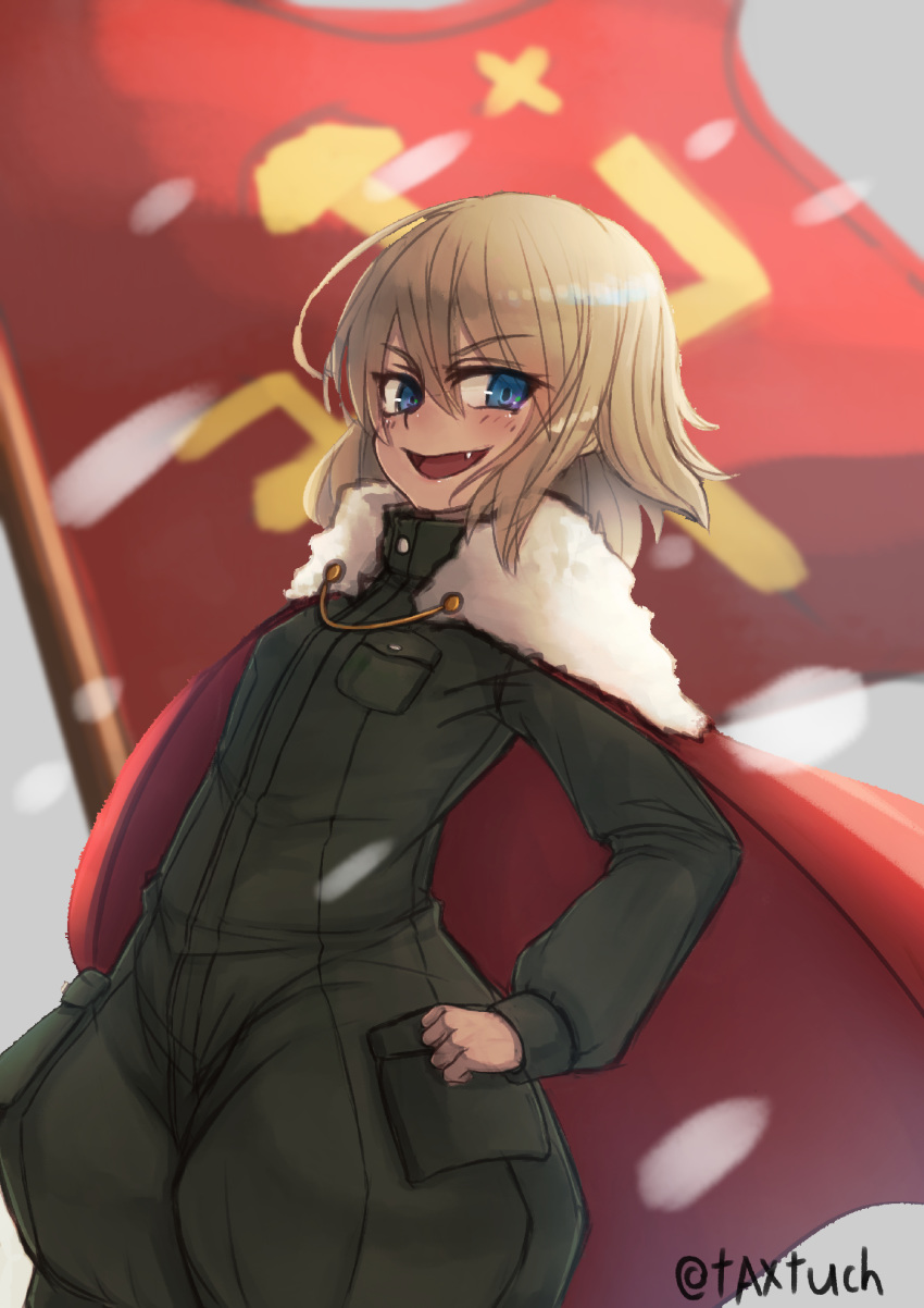 1girl bangs blonde_hair blue_eyes blurry blurry_background cape commentary emblem eyebrows_visible_through_hair fang flag girls_und_panzer green_jumpsuit hand_on_hip highres katyusha light_blush long_sleeves looking_at_viewer military military_uniform open_mouth pravda_(emblem) pravda_military_uniform red_cape red_flag short_hair short_jumpsuit smirk solo standing tacch twitter_username uniform