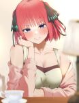  1girl bare_shoulders blue_eyes blush breasts butterfly_hair_ornament cardigan cleavage clenched_teeth cup elbow_rest go-toubun_no_hanayome green_shirt hair_ornament hand_in_own_hair head_rest highres indoors kakato_0 lamp large_breasts long_sleeves looking_at_viewer nakano_nino off_shoulder open_cardigan open_clothes pink_cardigan pink_hair shirt short_hair smile solo t-shirt table teacup teeth upper_body 