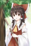 1girl ascot bangs bare_shoulders blurry blurry_background blush broom brown_eyes brown_hair collared_shirt commentary_request detached_sleeves frilled_shirt_collar frills hair_between_eyes hakurei_reimu hands_up highres holding holding_broom long_sleeves looking_at_viewer medium_hair open_mouth outdoors red_shirt red_skirt reimu_tyuki shirt skirt skirt_set sky solo standing sunlight touhou tree white_sky wide_sleeves yellow_ascot 