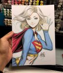  1girl blonde_hair blue_eyes blue_outline breasts cape dated dc_comics from_side hand_up highres long_sleeves looking_at_viewer medium_breasts midriff navel photo red_cape signature smile solo stanley_lau supergirl traditional_media waving white_background 
