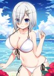  1girl beach bikini blue_eyes breasts collarbone eyebrows_visible_through_hair eyes_visible_through_hair flower front-tie_top hair_ornament hair_over_one_eye hairclip hamakaze_(kantai_collection) hibiscus highres kantai_collection kuroko_(user_jtty5588) large_breasts looking_at_viewer navel o-ring o-ring_bikini o-ring_top ocean short_hair side-tie_bikini silver_hair solo swimsuit water white_bikini 