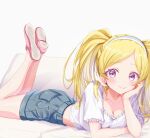  1girl absurdres bare_legs blonde_hair blush breasts cleavage collarbone cropped_shirt denim denim_shorts emily_stewart feet_up hand_on_own_cheek hand_on_own_face head_rest highres idolmaster idolmaster_million_live! looking_at_viewer lying on_stomach purple_eyes saki_nerikeshi shorts small_breasts smile solo the_pose twintails 