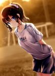  1girl absurdres arms_behind_back blue_eyes blurry blurry_background breasts brown_hair chikasugiru_karera_no_juunanasai_no_tooi_kankei closed_mouth clothes_writing collarbone dutch_angle evening highres kazuharu_kina leaning_forward long_hair looking_at_viewer medium_breasts mori_yuriko novel_illustration official_art outdoors pink_scrunchie ponytail scrunchie shirt short_sleeves shorts sidelocks smile soccer_uniform solo sportswear standing sunset wavy_hair white_shirt 