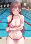  1girl bikini blurry breasts brown_hair cleavage collarbone commentary_request cowboy_shot depth_of_field highres indoors kanojo_okarishimasu large_breasts leaning_forward long_hair looking_at_viewer mizuhara_chizuru navel pink_bikini pool red_eyes side-tie_bikini solar_(happymonk) solo swimsuit 