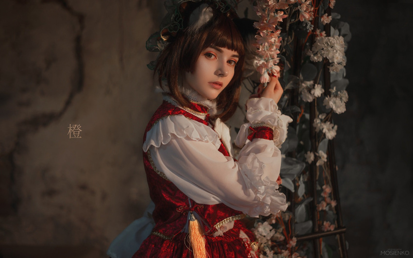 1girl animal_ears blurry blurry_background brown_eyes brown_hair cat_ears chen commentary cosplay cosplay_photo cowboy_shot depth_of_field embellished_costume english_commentary flower from_side highres lips long_sleeves looking_at_viewer looking_to_the_side makeup photo_(medium) pink_flower red_skirt red_vest shihori_chan shirt skirt skirt_set solo tassel touhou vest white_flower white_shirt