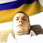  brown_hair closed_mouth indoors looking_at_viewer non-web_source off-topic photo_(medium) short_hair ugly_man ukrainian_flag upper_body very_short_hair 