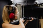  1girl bare_arms blonde_hair blurry ear_protection from_side gun holding holding_gun holding_weapon indoors looking_ahead off-topic outdoors photo_(medium) pointing_gun pump_action remington_870 shooting_glasses shooting_range short_hair shotgun solo upper_body watch weapon wristwatch 
