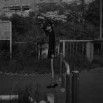  1girl absurdres arms_at_sides building bush commentary_request fence from_side greyscale highres long_hair long_sleeves looking_at_viewer looking_to_the_side monochrome narue original outdoors photo_background sign socks solo standing 