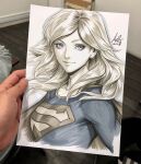  1girl artist's_hand_in_frame blonde_hair blue_eyes blue_shirt cape closed_mouth commentary cropped_torso dc_comics english_commentary highres long_hair looking_at_viewer marker_(medium) photo_(medium) shirt signature solo stanley_lau supergirl superman_(series) superman_logo traditional_media turning_head 
