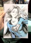  1girl art_tools_in_frame artist's_hand_in_frame blue_eyes blue_shirt breasts cape closed_mouth collarbone commentary dc_comics english_commentary heart highres long_hair looking_at_viewer marker_(medium) one_eye_closed photo_(medium) shirt signature solo stanley_lau supergirl superman_(series) superman_logo traditional_media v white_background 
