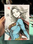  1girl art_tools_in_frame artist's_hand_in_frame blonde_hair blue_eyes breasts cape clenched_hand closed_mouth commentary dc_comics english_commentary heart highres large_breasts long_hair looking_at_viewer marker_(medium) photo_(medium) red_cape red_skirt signature skirt stanley_lau supergirl superhero_costume superman_(series) superman_logo traditional_media turning_head upper_body 