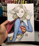 1girl art_tools_in_frame artist's_hand_in_frame blonde_hair blue_eyes blue_outline breasts cape closed_mouth commentary crop_top dc_comics english_commentary from_side hand_up highres long_sleeves looking_at_viewer marker_(medium) medium_breasts medium_hair midriff navel outline photo_(medium) red_cape signature smile solo stanley_lau supergirl supergirl_(2004_costume) superman_(series) traditional_media waving white_background 