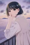 1girl bangs black_hair blurry blurry_background blush brown_dress closed_mouth depth_of_field dress eyebrows_behind_hair frilled_shirt frilled_sleeves frills from_side grey_eyes hand_to_own_mouth hand_up long_sleeves looking_at_viewer looking_to_the_side mattaku_mousuke original outdoors pleated_dress railing shirt sleeveless sleeveless_dress smile solo sunset thick_eyebrows twitter_username water watermark white_shirt 