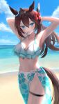  1girl ahoge animal_ears arm_up armpits bangs bare_shoulders beach bikini blue_bikini blue_sky breasts brown_hair cleavage cloud cowboy_shot flower frilled_bikini frills hair_between_eyes hair_flower hair_ornament hair_tie_in_mouth hairband hibiscus hibiscus_print highres horse_ears horse_girl horse_tail large_breasts long_hair m.a.y. mihono_bourbon_(umamusume) mouth_hold navel ocean outdoors sand sarong sidelocks sky solo stomach swimsuit tail thigh_strap thighs tying_hair umamusume wet 