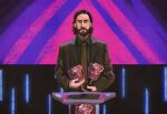  1boy alan_wake alan_wake_(game) beard black_hair blue_eyes brown_coat brown_necktie coat collared_shirt facial_hair game_show green_shirt holding holding_trophy looking_at_viewer male_focus medium_hair necktie neo_jpeg neon_lights podium shirt solo surprised the_game_awards trophy upper_body wide-eyed 