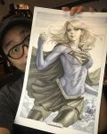  1girl artist's_hand_in_frame blonde_hair blue_shirt breasts cape closed_mouth commentary dc_comics english_commentary flying highres light_smile long_hair long_sleeves marker_(medium) photo_(medium) sepia shirt signature skirt solo stanley_lau supergirl superman_(series) superman_logo traditional_media 