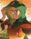 1girl amphibia armor autumn_leaves black_shirt blonde_hair blurry blurry_background clenched_teeth falling_leaves grass green_eyes green_hood highres hood hood_up jocy3 leaf leaf_on_head looking_at_viewer mole mole_under_eye outdoors red_armor sasha_waybright scale_armor shirt short_sleeves smile teeth turtleneck upper_body wood 